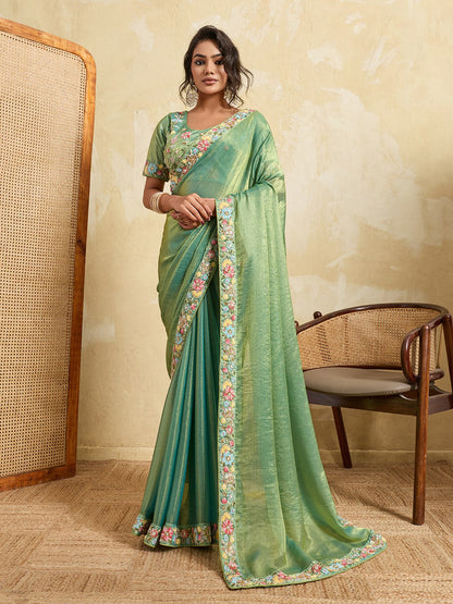 Woman wearing a green saree with floral patterns in a room with a chair and woven wall.